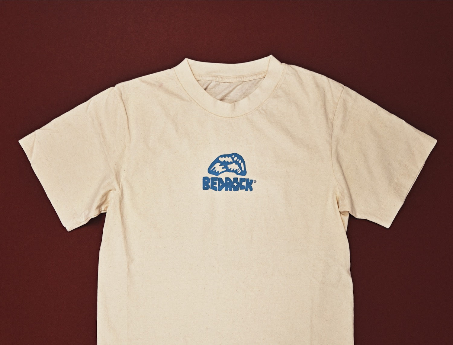 A beige crew neck T-shirt with the brand name 'BEDROCK' printed in blue on the chest.