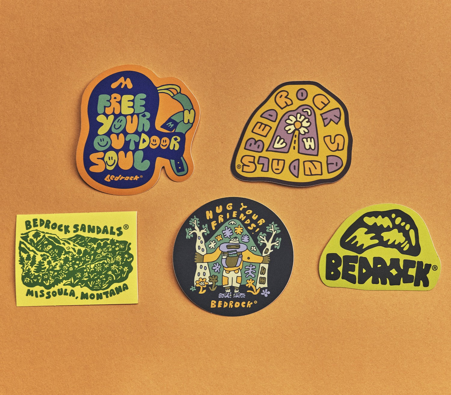 A collection of colorful Bedrock die-cut stickers with various designs, including text and the Bedrock brand logo, placed on a brown surface.