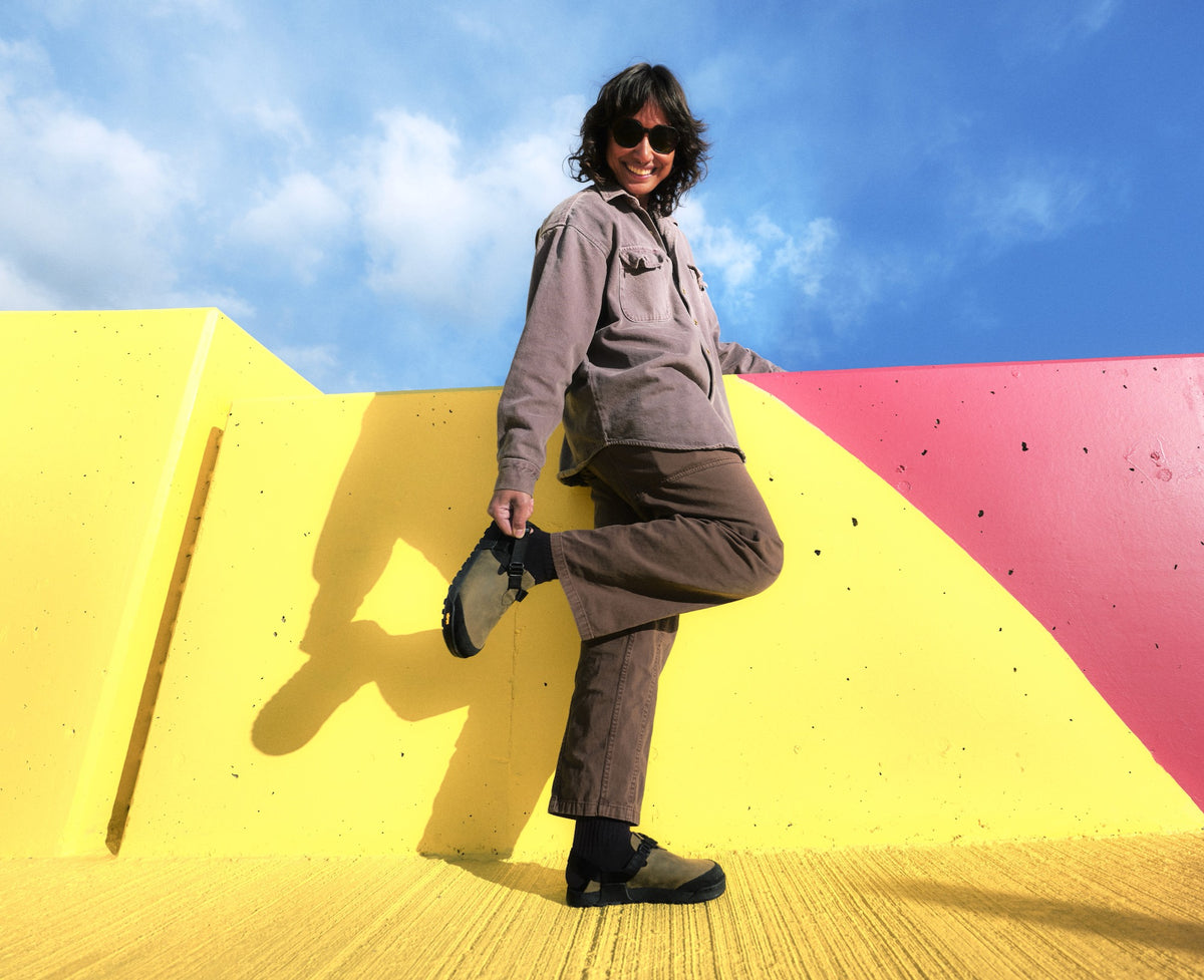 A Person smiling and posing in a brightly colored yellow urban backdrop wearing Driftwood gray Nubuck leather Mountain Clogs