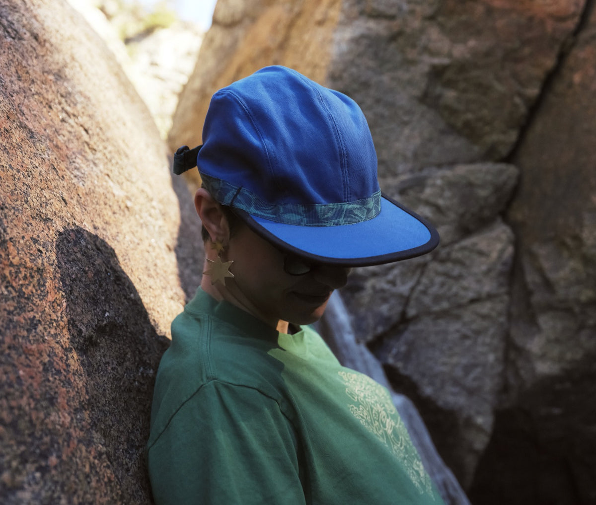 A person looking at the ground wearing a blue organic cotton canvas fabric hat with River Mosaic patterned webbing by KAVU and Bedrock.