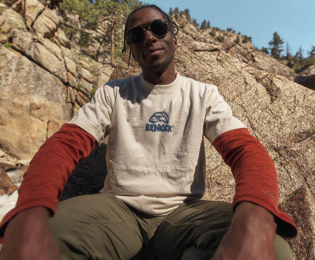 A person looking at the camera while sitting on granite rock wearing A beige crew neck T-shirt with the brand name 'BEDROCK' printed in blue on the chest.