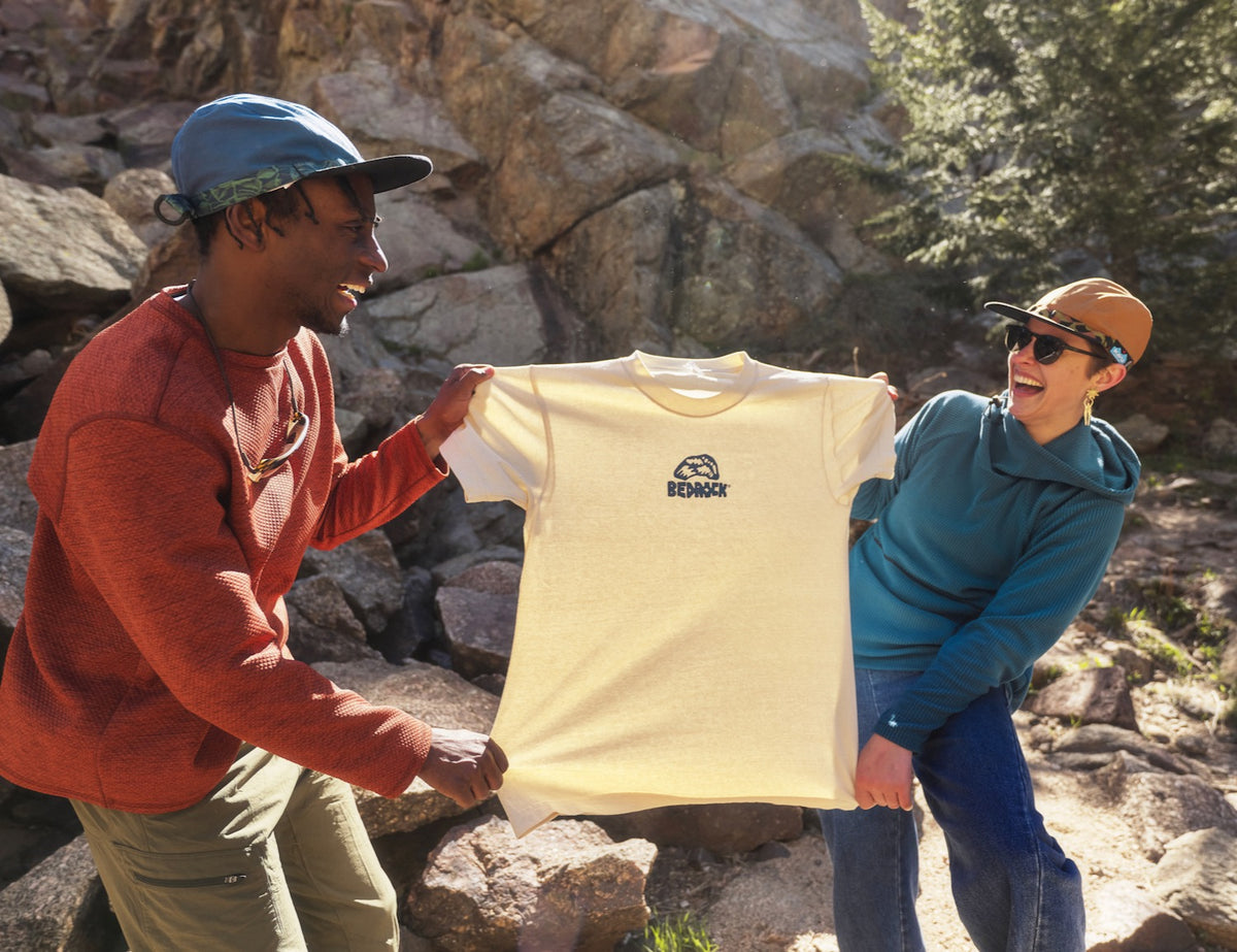 Two people smiling and holding a beige crew neck T-shirt with the brand name 'BEDROCK' printed in blue on the chest.