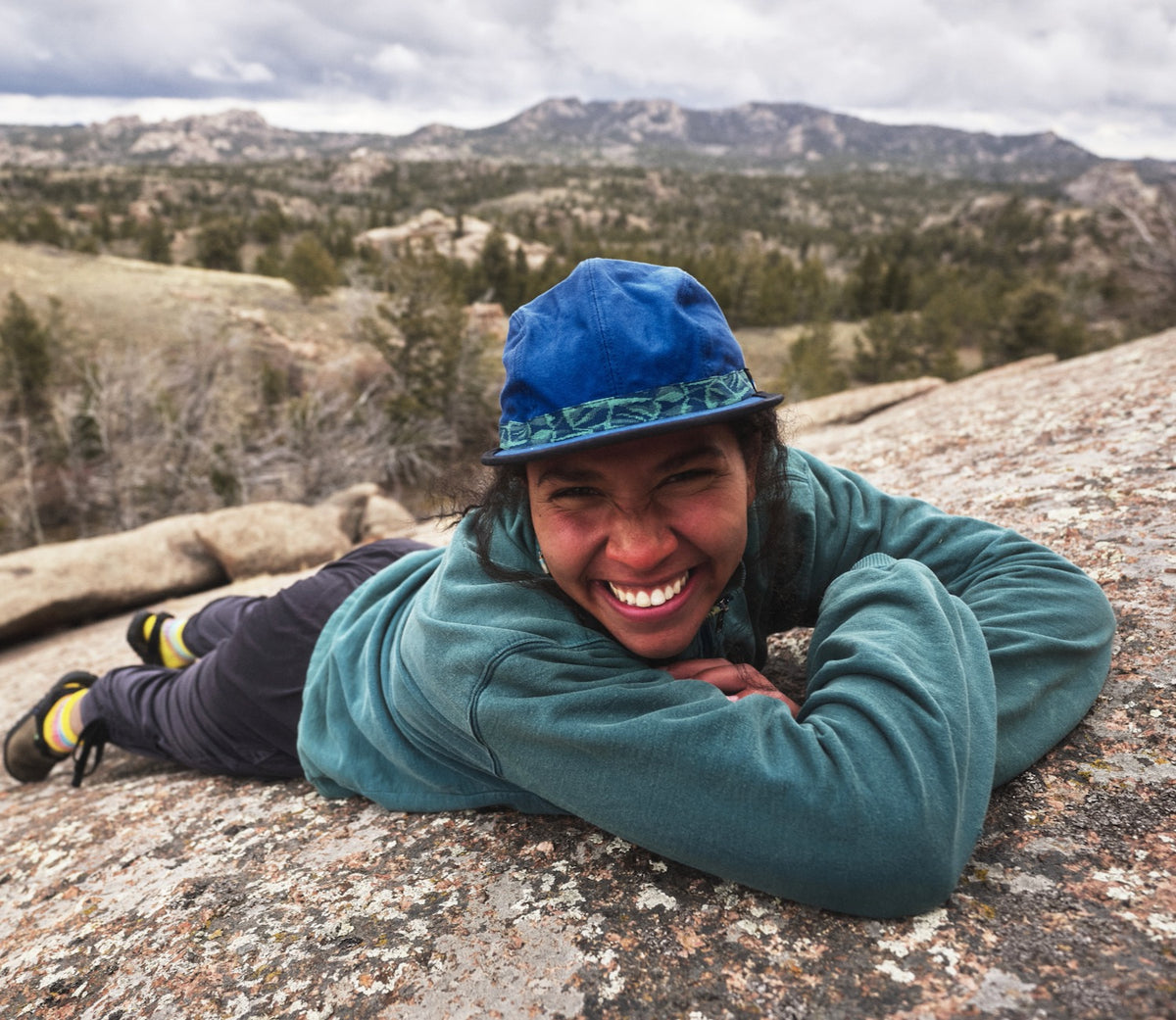 A person smiling at camera laying on granite rock, wearing a blue organic cotton canvas fabric hat with River Mosaic patterned webbing by KAVU and Bedrock.
