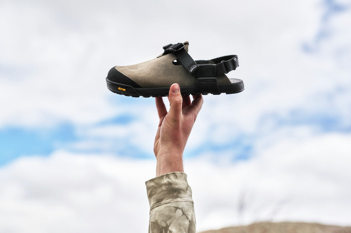 A hand holding up a pair of Driftwood gray Nubuck leather Mountain Clogs with a blue sky backdrop