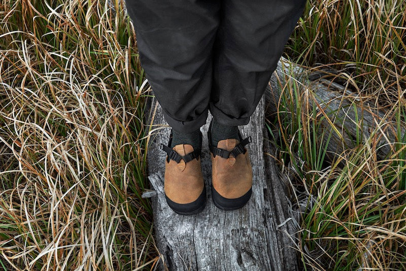 person wearing brown nubuck leather mountain clogs on a log amidst grass