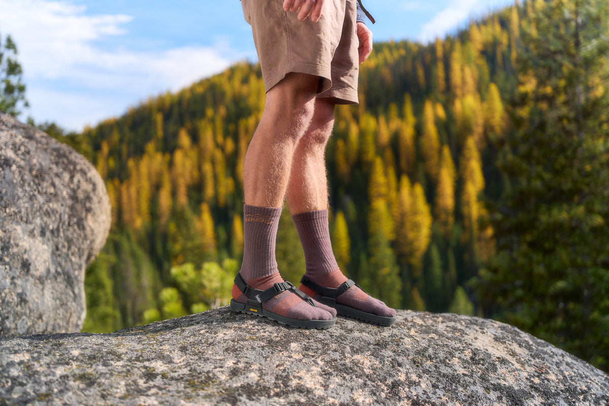 Person standing on a rock with a forest and blue sky in the background. Wearing Pair of Crew length plum split-toe socks