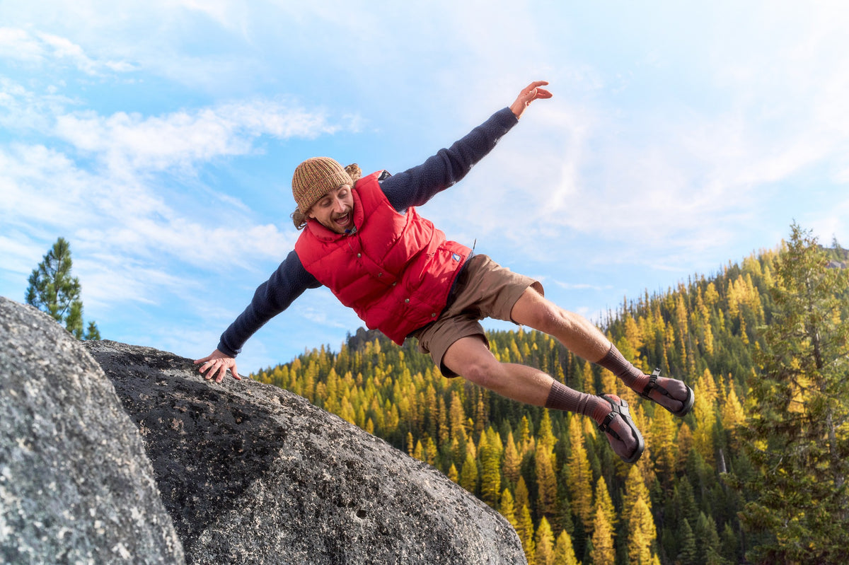 Person in red vest and tan hat climbing a rock with a mountainous landscape in the background.