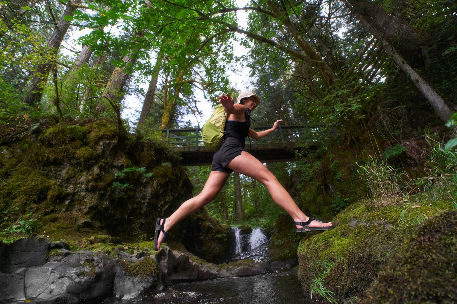 Person in a black outfit and hat jumping over a stream in a forest wearing Cairn Evo C PRO sandals