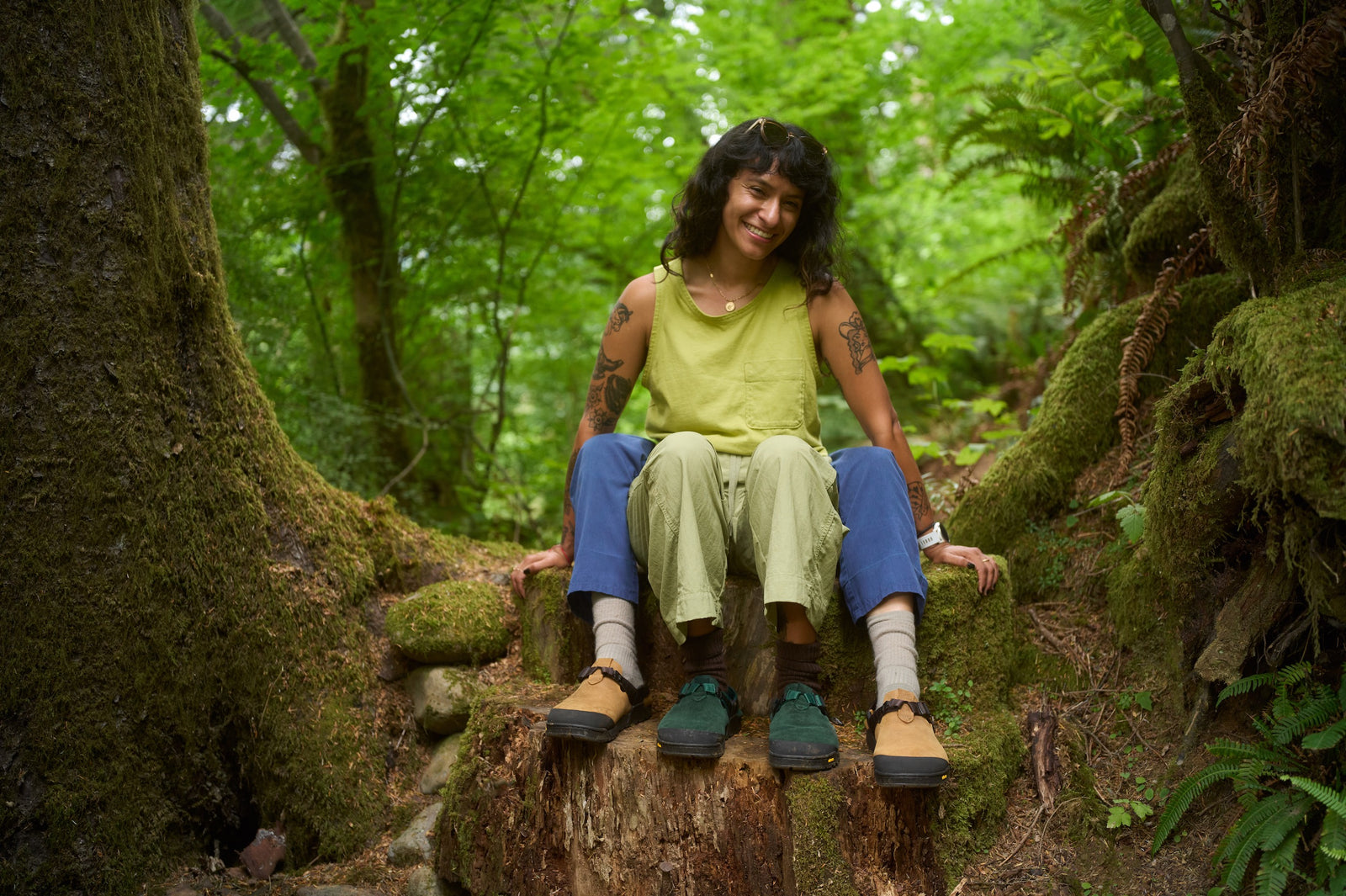 Two people sitting on a log in a forest highlighting their Mountain Clogs in Cascade Green Leather Suede and in Nubuck Brown