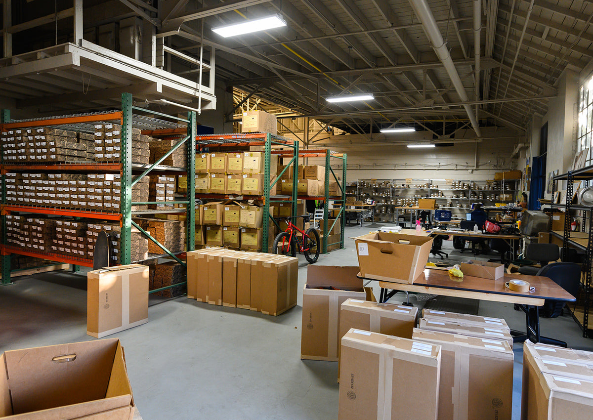 A large warehouse interior with tall shelving units filled with boxes, cardboard pallets, work tables, and equipment under bright overhead fluorescent lights.