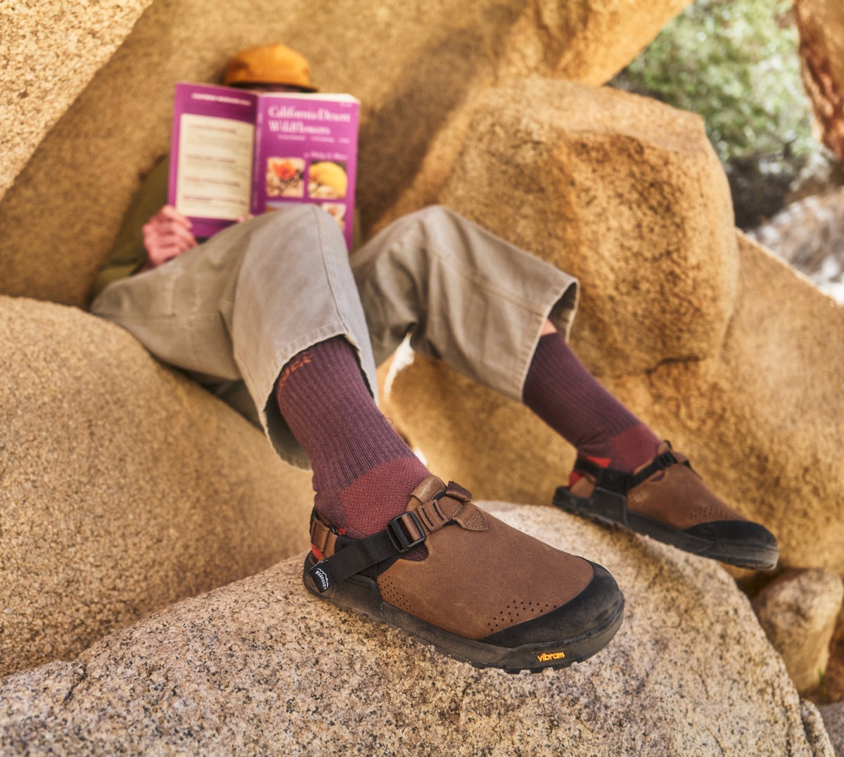 Person wearing Pinecone Synthetic Suede Mountain Clogs and purple socks sitting on rocks with a book.