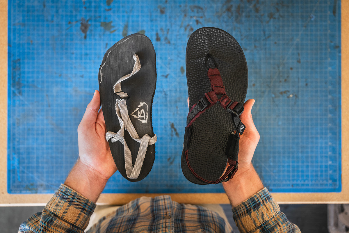 A top-down view of a person holding two sandals over a blue gridded work surface. The left sandal is black with light gray straps and a white logo, and the right sandal is black with dark red straps and a textured sole.