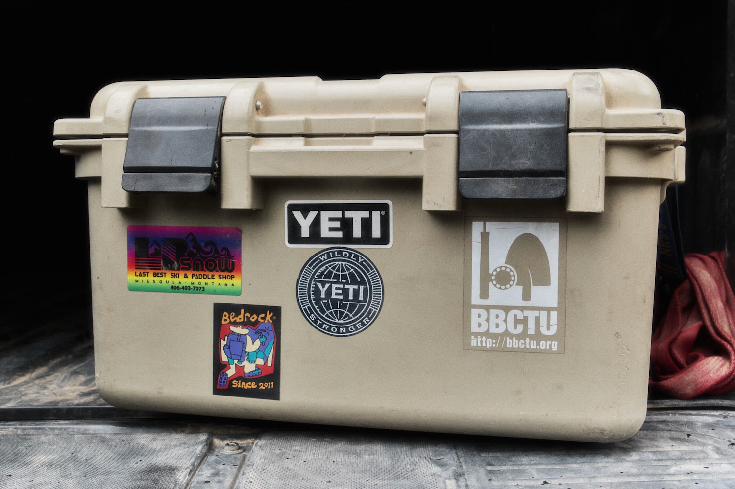 A beige YETI hard-sided cooler or storage box with black latches and several stickers on the front, including YETI branding and BBCTU, placed on a wooden surface against a dark background.