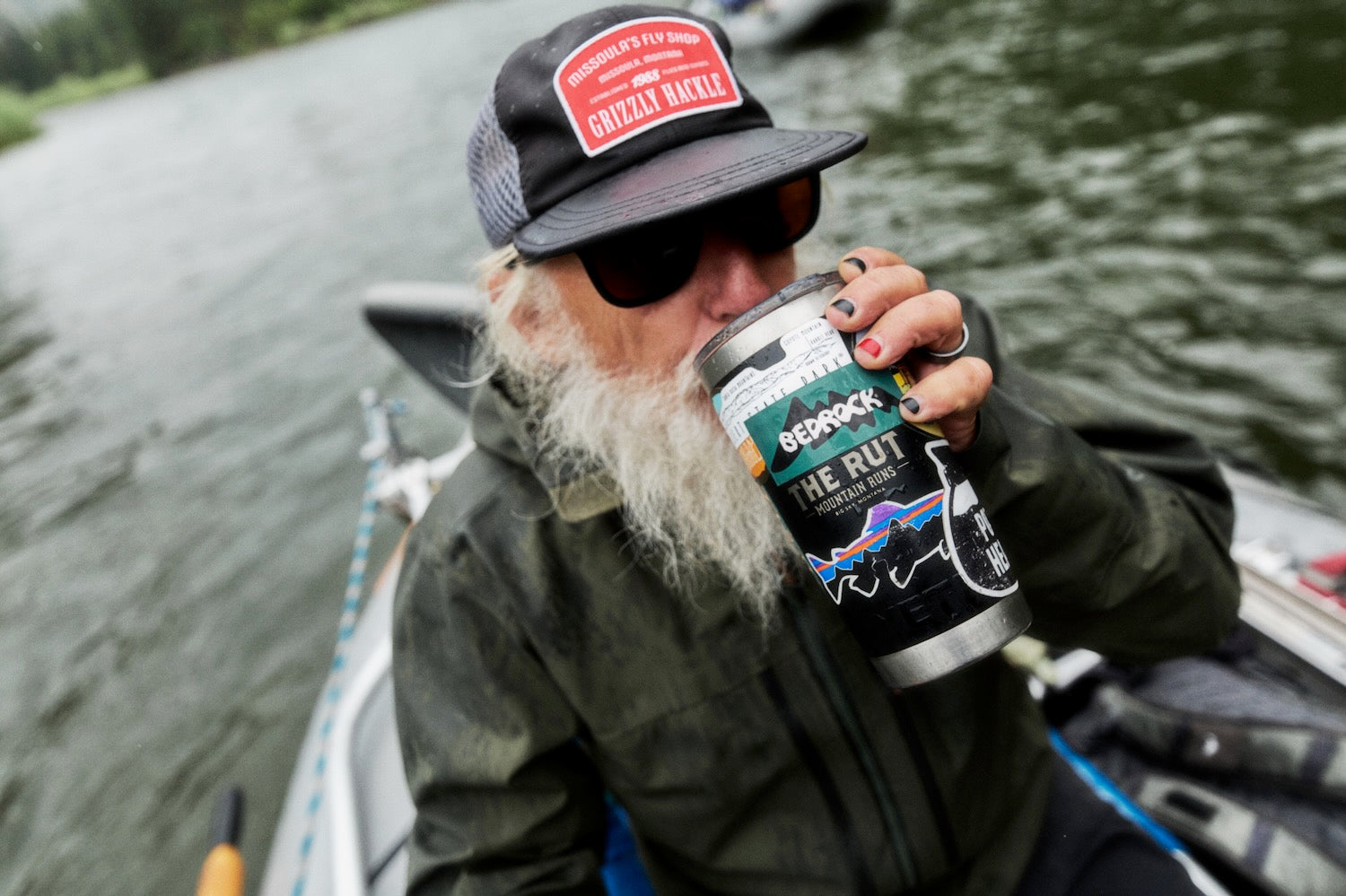 A bearded man wearing a black cap with a red 'Grizzly Barrel' logo and sunglasses, sitting in a boat on the water and drinking from a waterbottle.