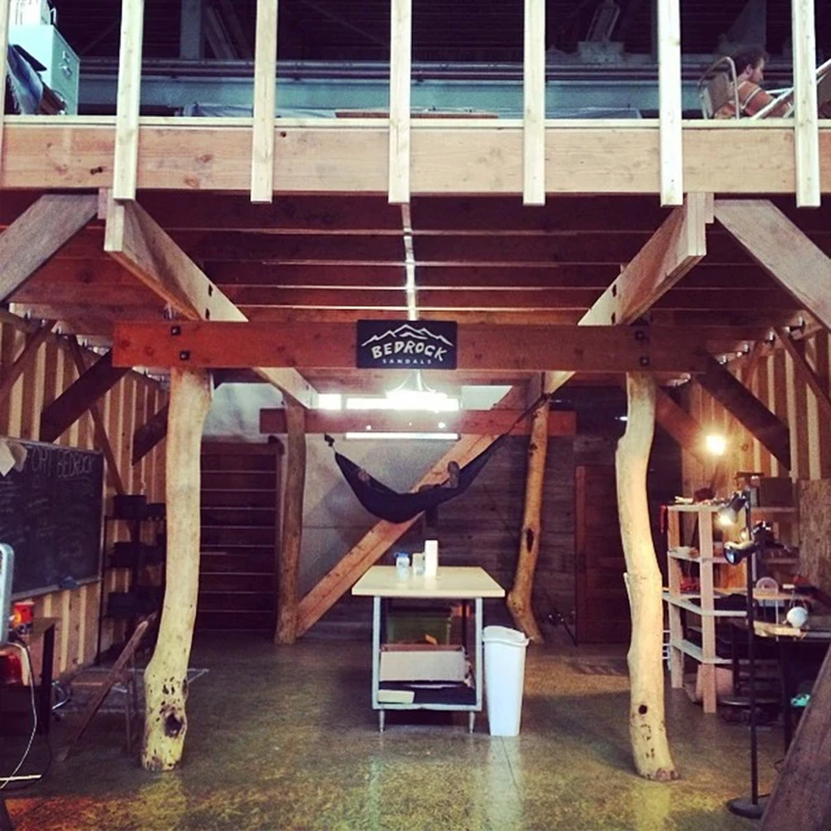 A rustic loft-style wooden cabin interior with a loft floor, exposed beams, a hanging hammock, a small table in the center, and a sign reading 'BEPROOZ' hanging from the ceiling.