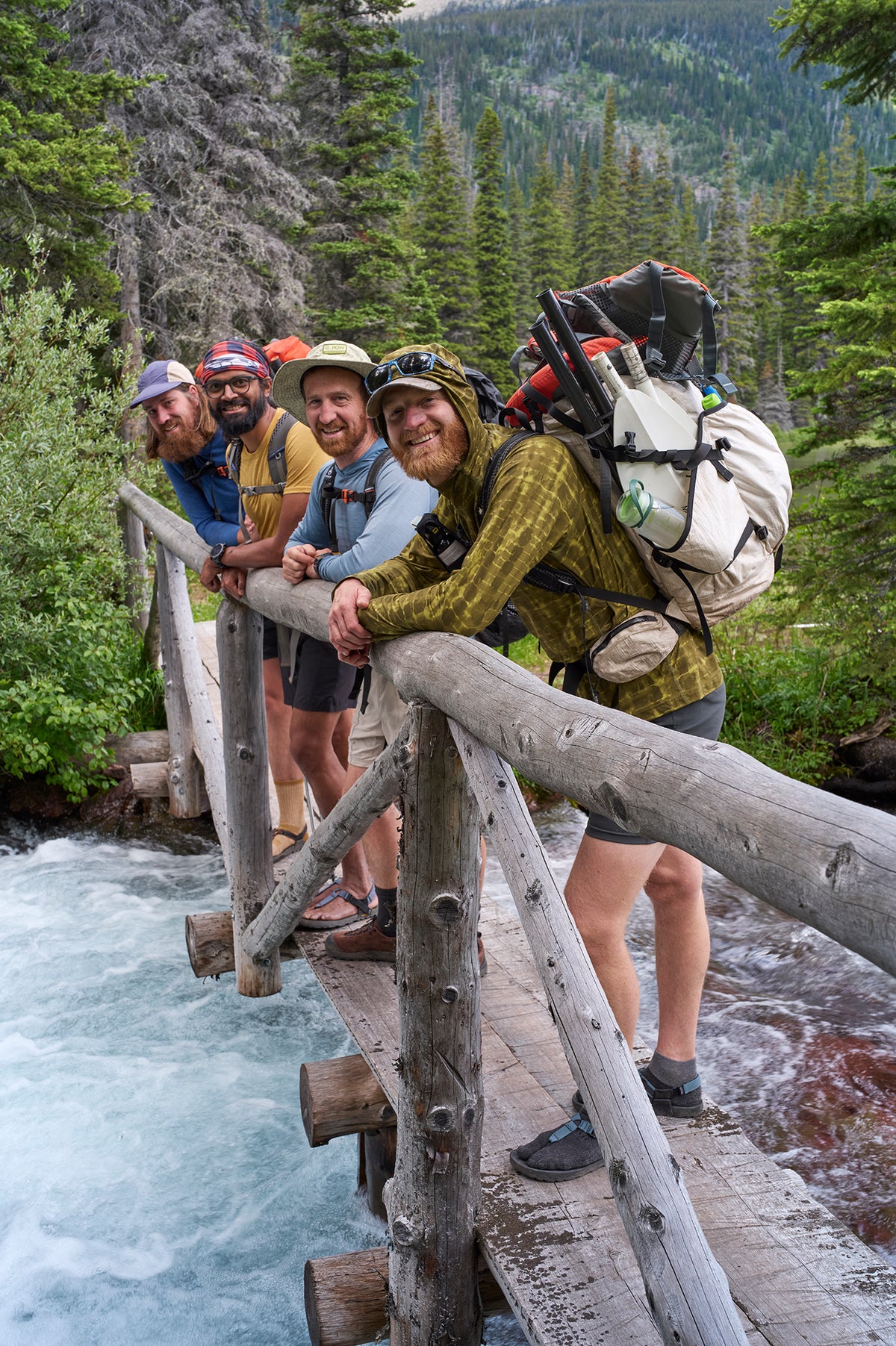 Four hikers with large backpacks stand on a rustic wooden bridge over a rushing river, with a forested mountain in the background.