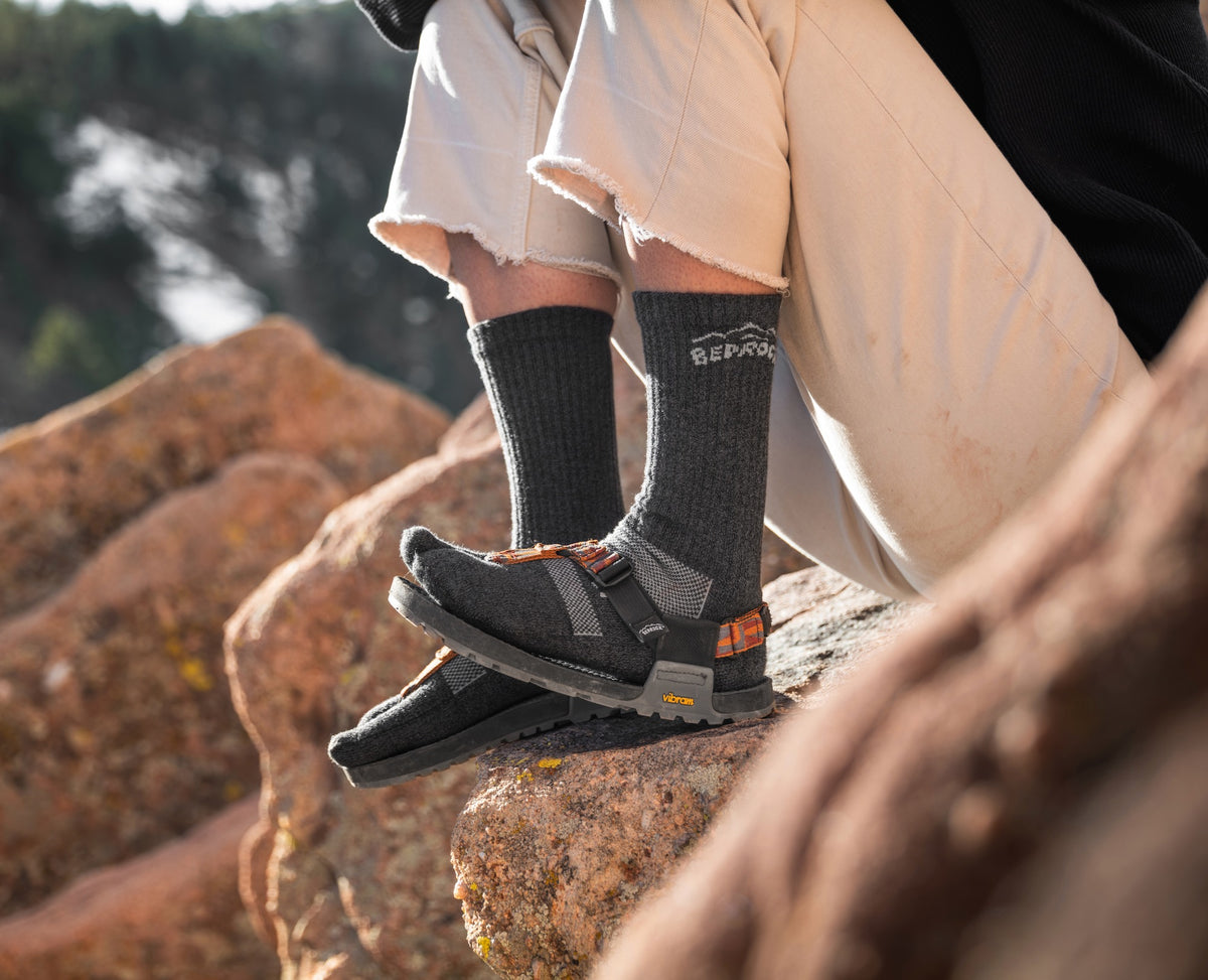 A person on a rockwearing a pair of cairn sandals with a pair of gray Bedrock x Injinji crew split-toe socks on, while flexing their toes up.