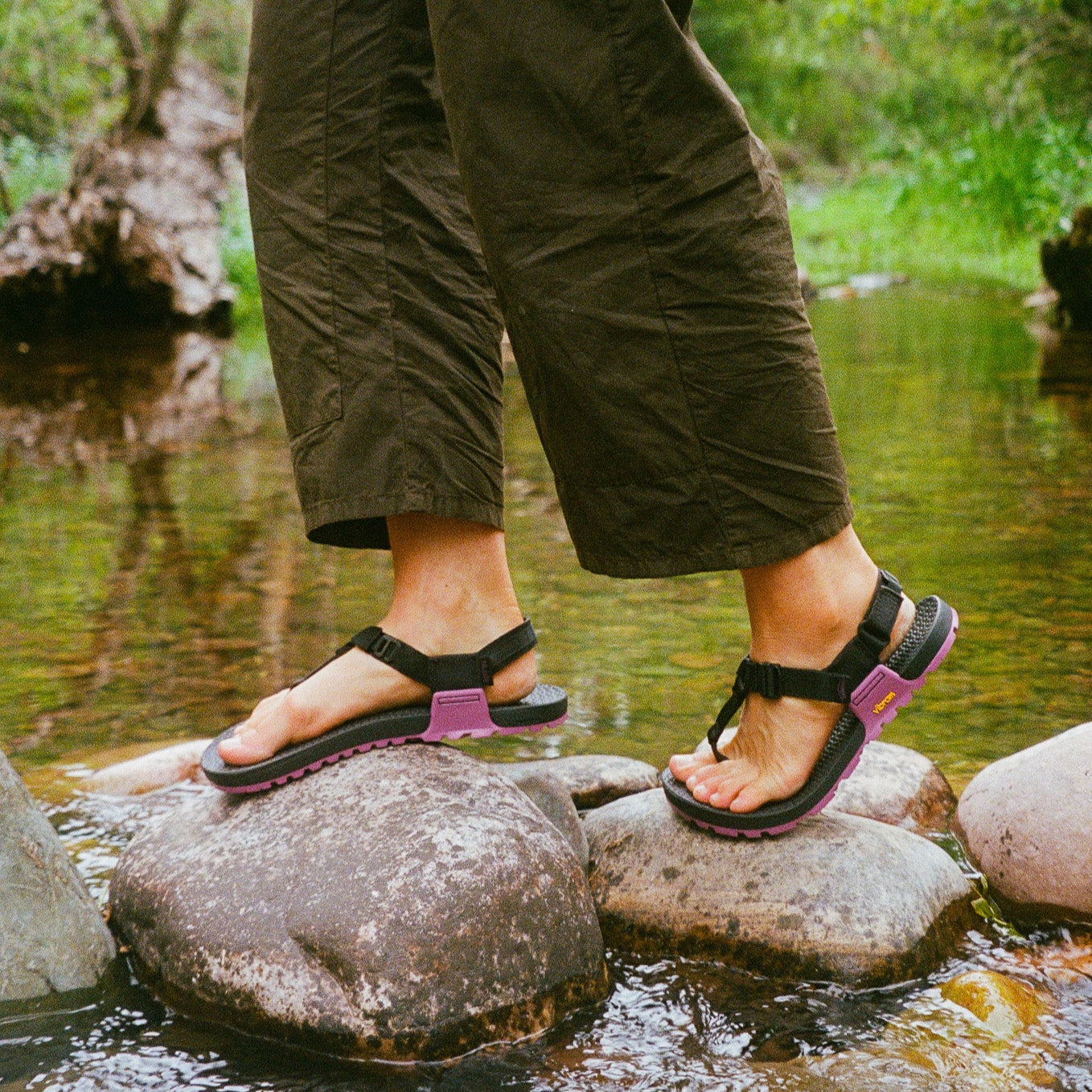 Person wearing sandals standing on rocks in a natural setting with greenery.