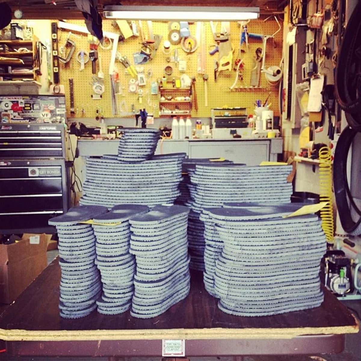 Stacks of gray textured circular discs, likely sanding or grinding wheels, arranged on a workbench in a cluttered workshop with tools and equipment in the background.