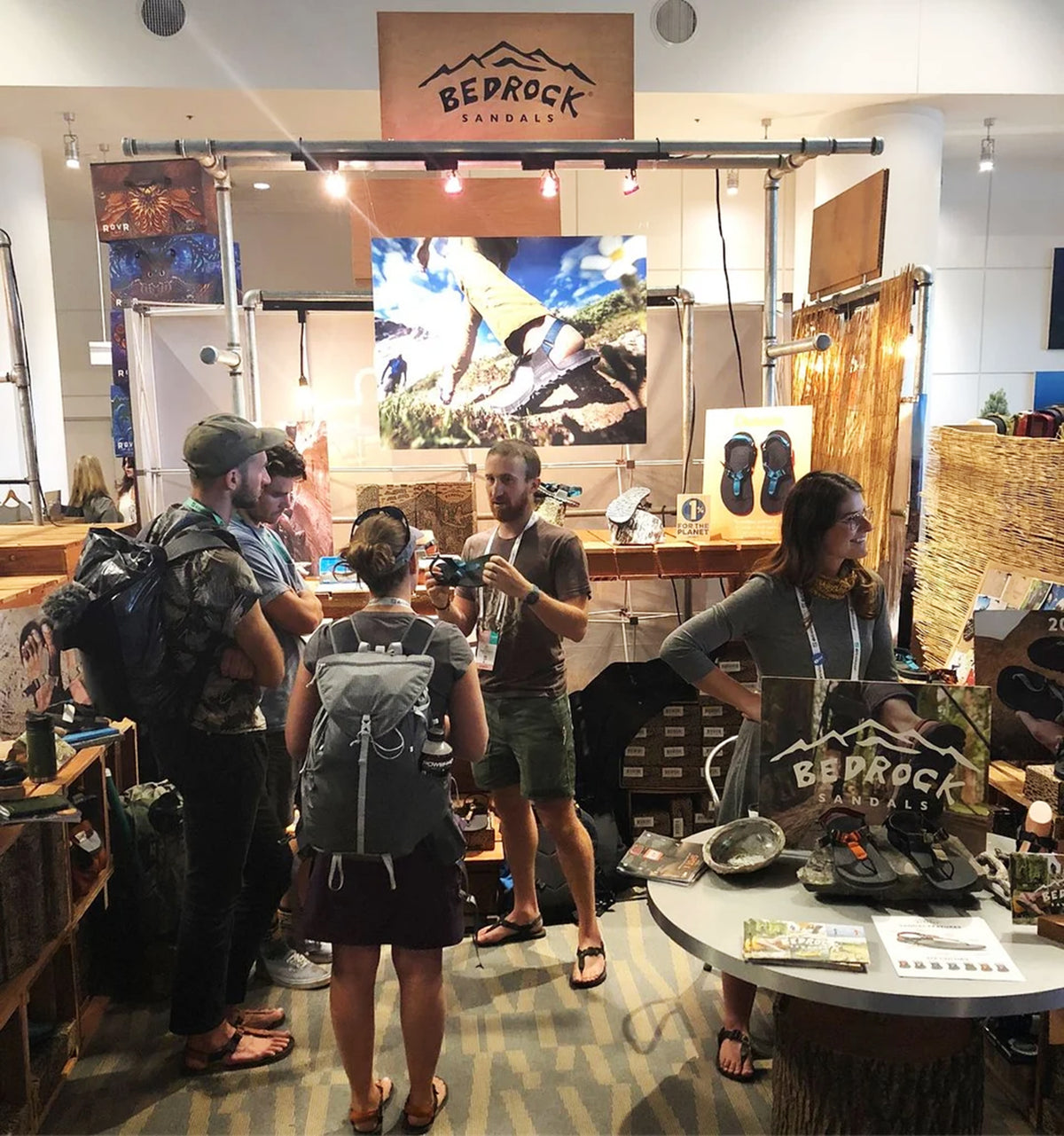 A Bedrock Outdoors booth at an indoor event, featuring a wooden sign, display of hiking gear, and several people interacting with staff behind a glass partition.