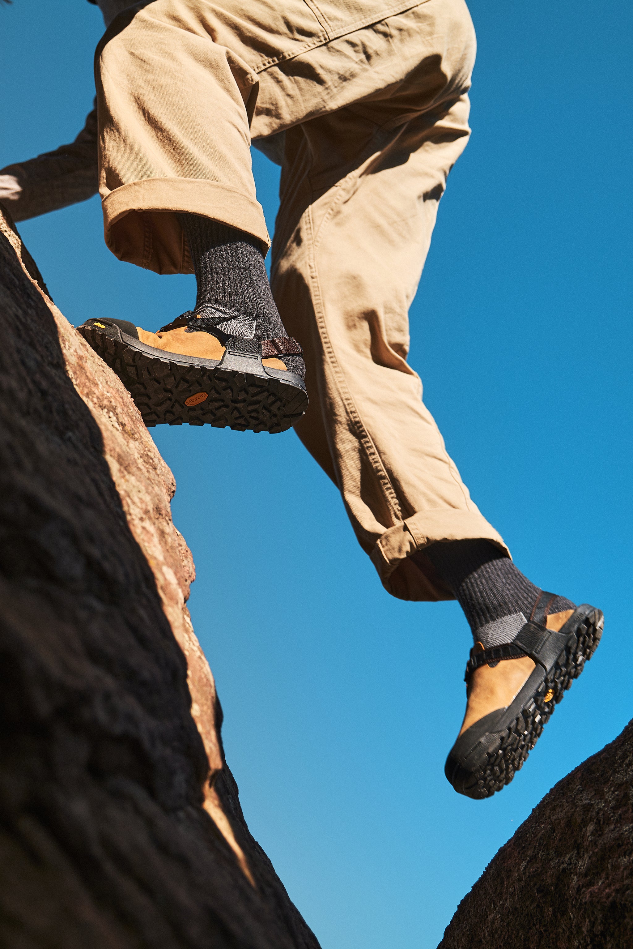 A person climbing a rocky outcrop, shown from below wearing tan pants, dark gray socks, and tan-and-black hiking shoes against a clear blue sky.