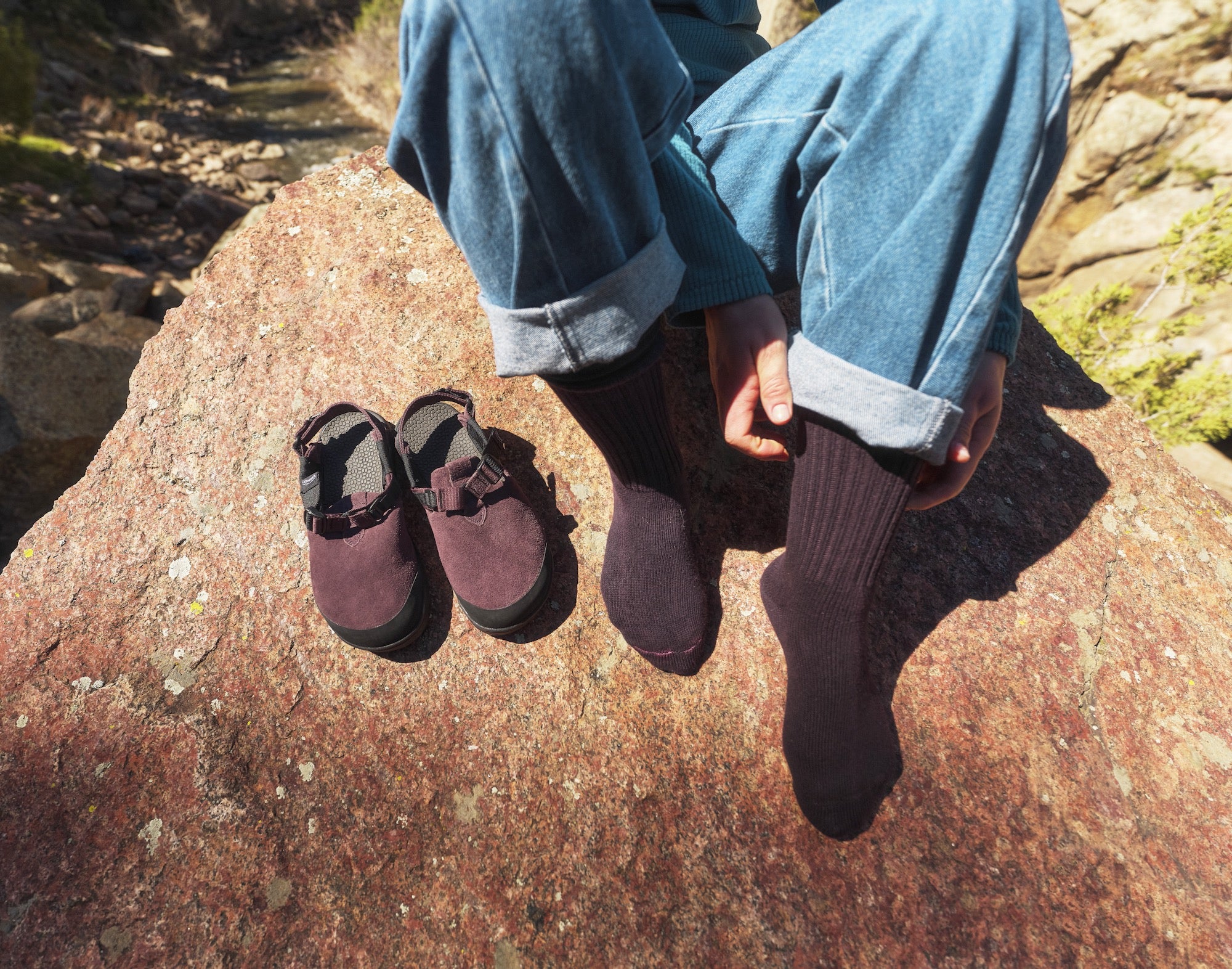 Person posing on a rock wearing purple socks about to put on Deep Plum Mountain Clogs