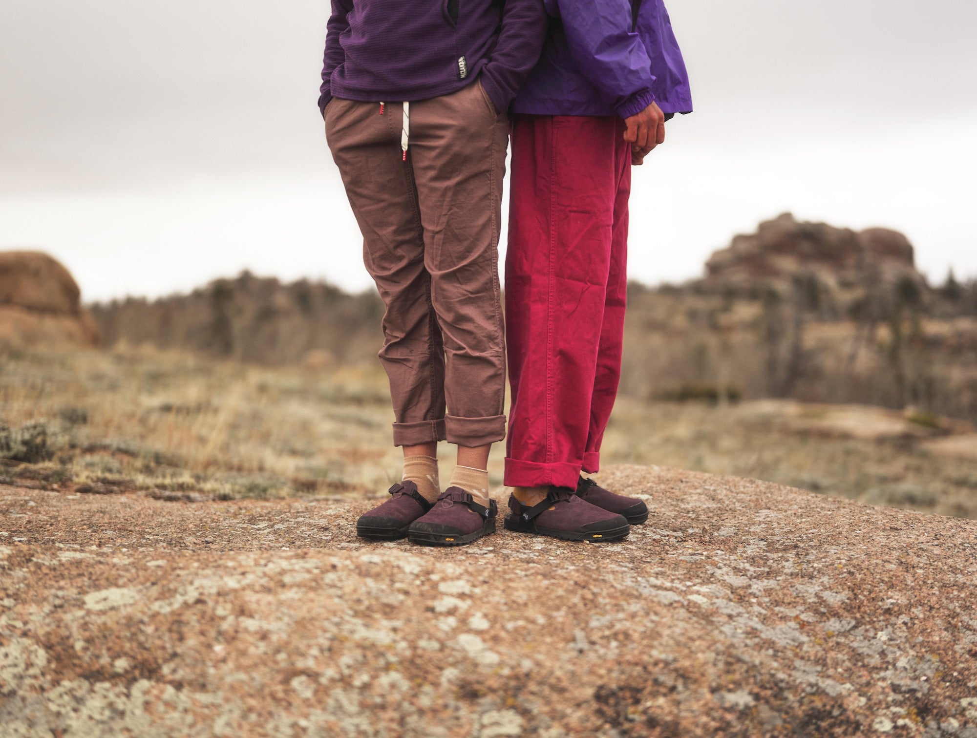 Two people posed back to back wearing Deep Plum Mountain Clogs in a mountain environment