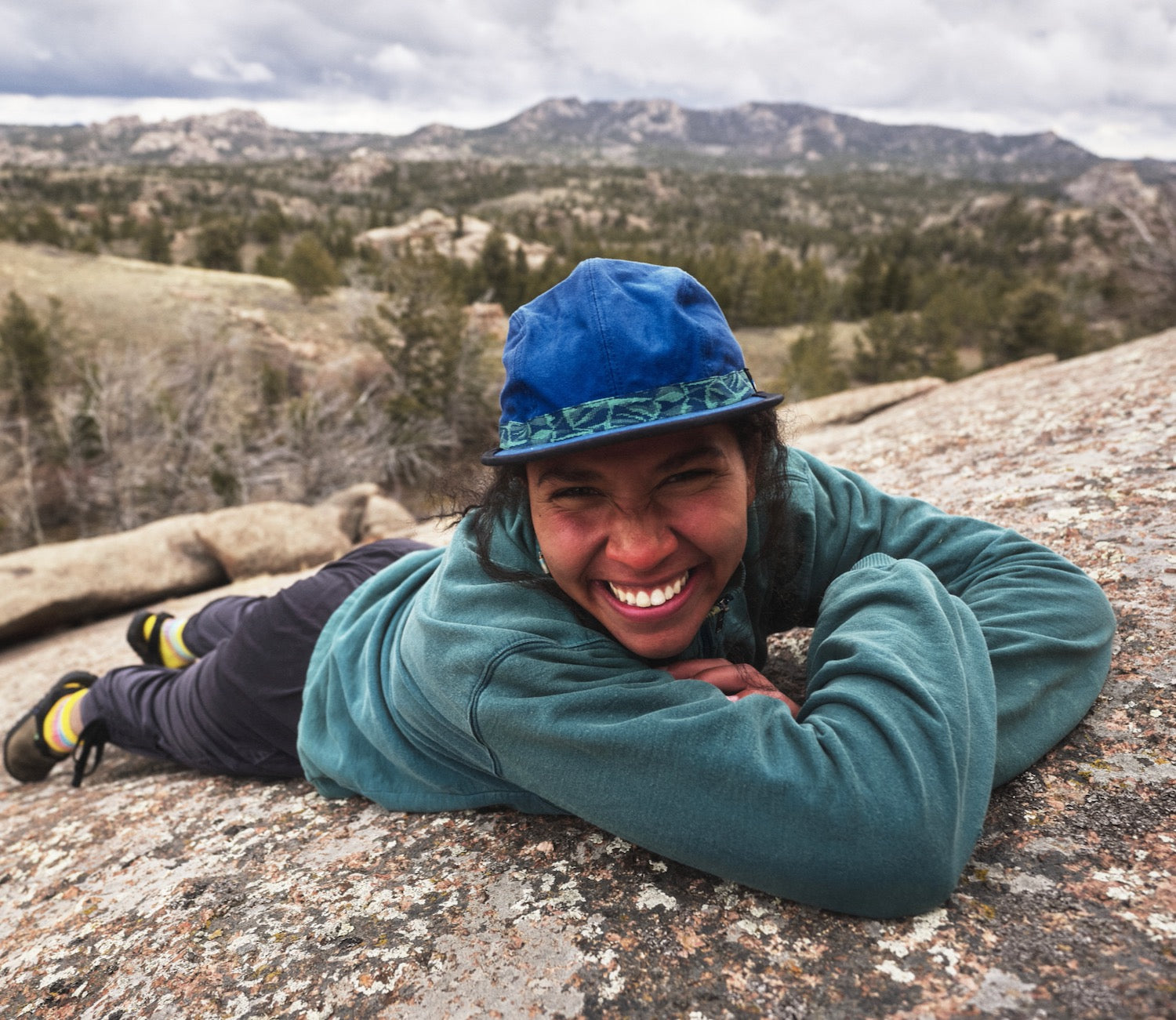A person smiling at camera laying on granite rock, wearing a blue organic cotton canvas fabric hat with River Mosaic patterned webbing by KAVU and Bedrock.