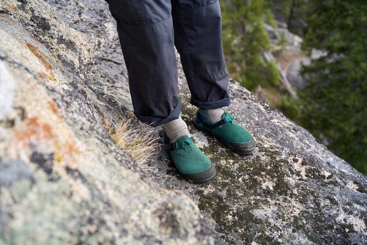 Person wearing Mountain Clogs in Cascade Green Leather Suede and dark pants standing on a rocky surface with a natural background.