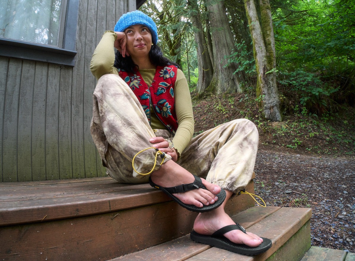 Person sitting on a wooden bench outdoors wearing a colorful outfit and Black Rockhound flip flops.