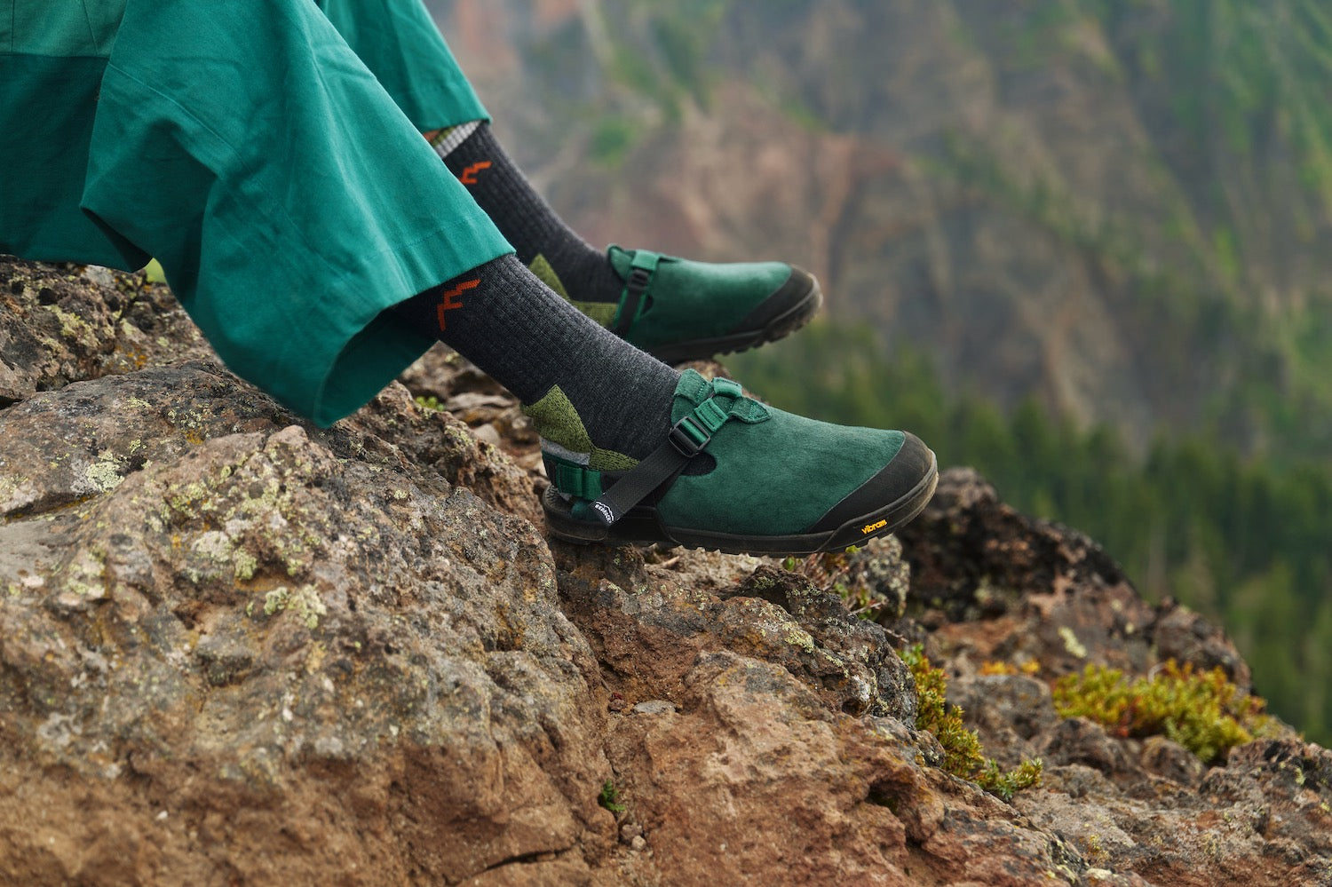 Person wearing Mountain Clogs in Cascade Green Leather Suede  and pants sitting on a rocky mountain ledge with a blurred natural background.