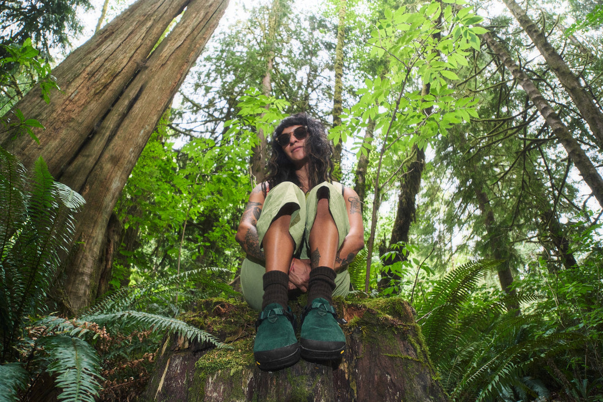 Person sitting on a log in a forest wearing Mountain Clogs in Cascade Green Leather Suede