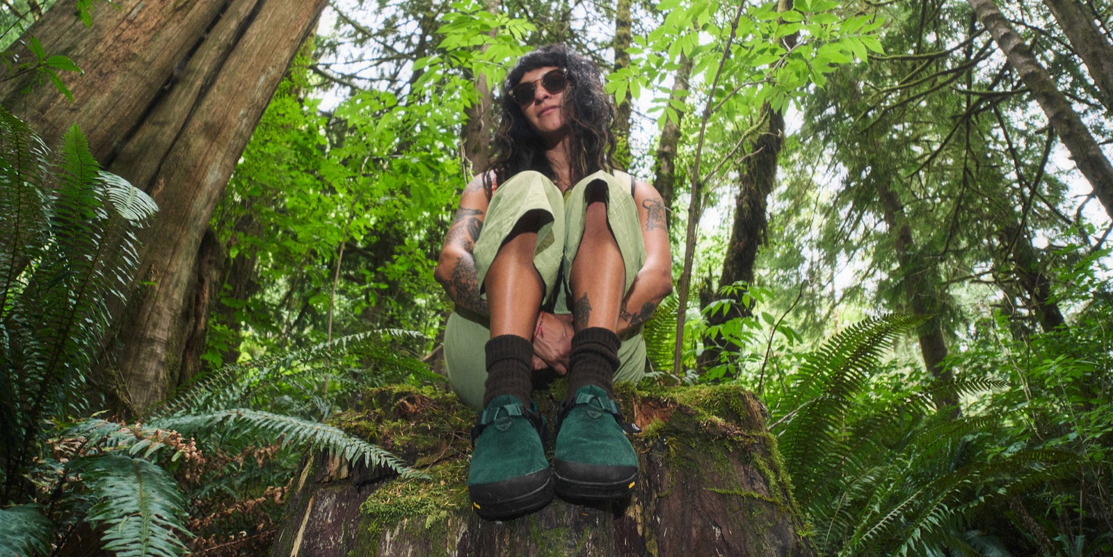 Person sitting on a log in a forest wearing cascade green mountain clogs and sunglasses.