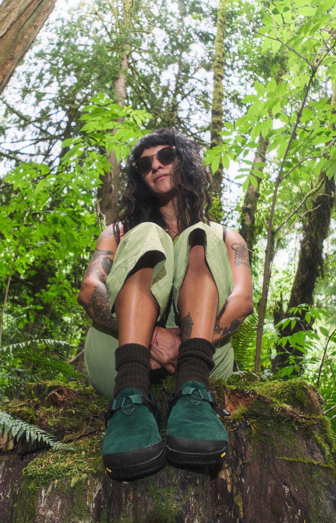 Person sitting on a log in a forest wearing cascade green mountain clogs and sunglasses.