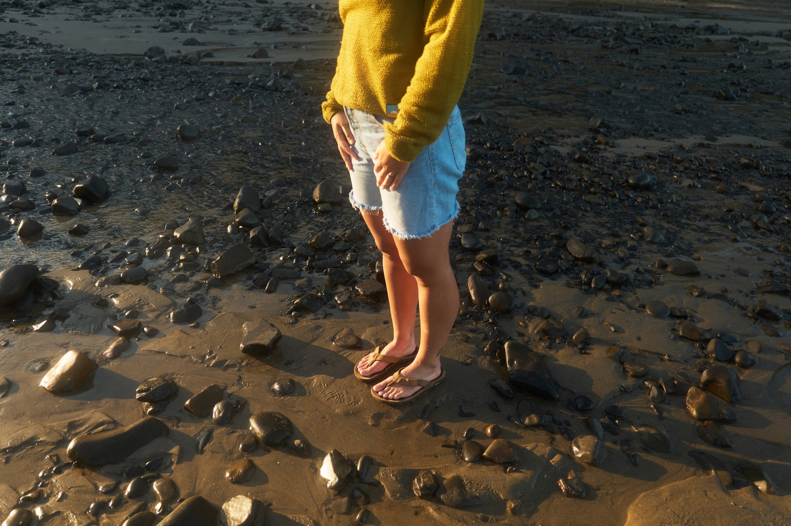 Person standing on a rocky beach wearing a yellow sweater, light blue shorts, and nubuck leather flip flops.