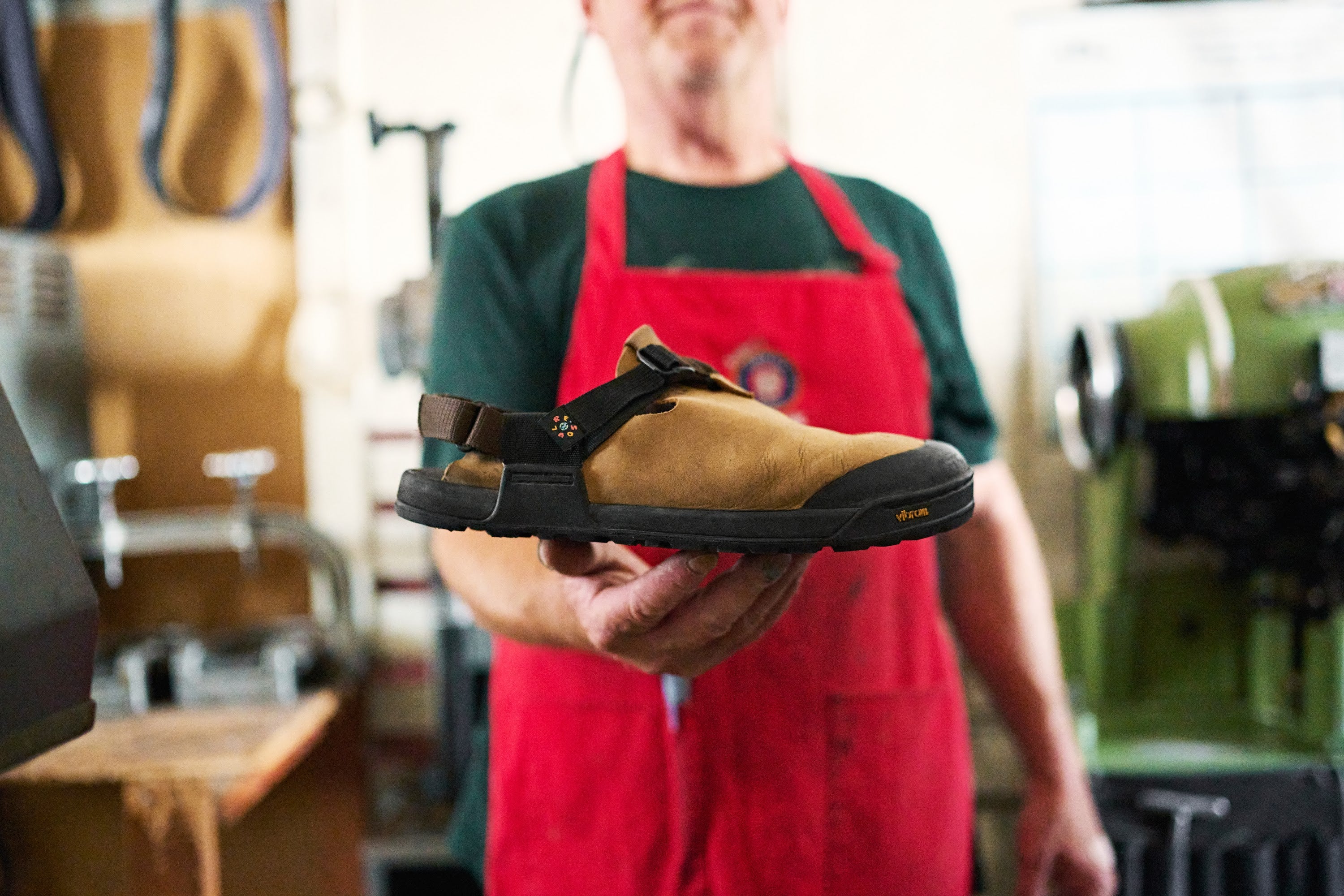 Person wearing a red apron holding a brown clog in a workshop setting