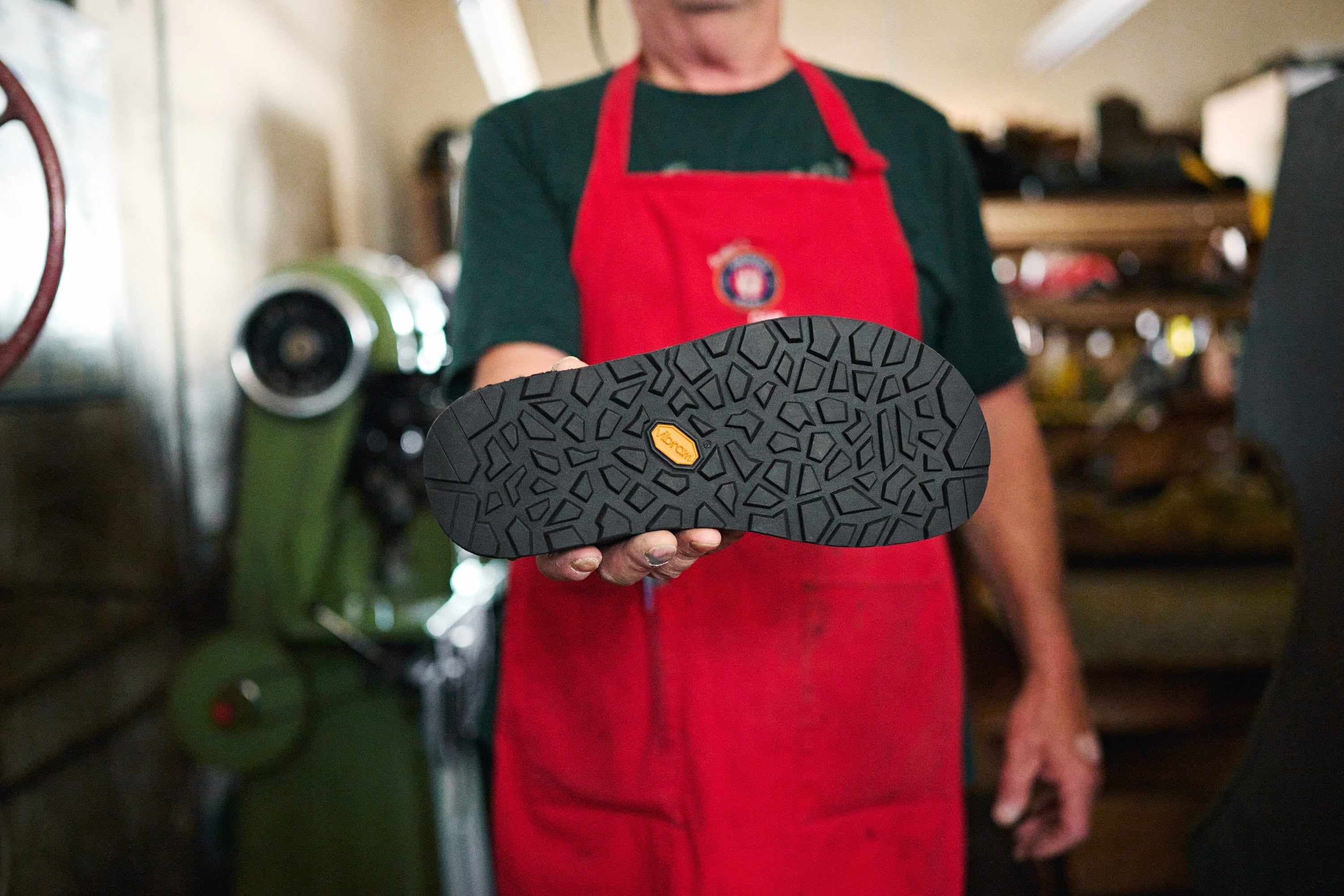 Person wearing a red apron holding a clog showing a Vibram sole in a workshop setting