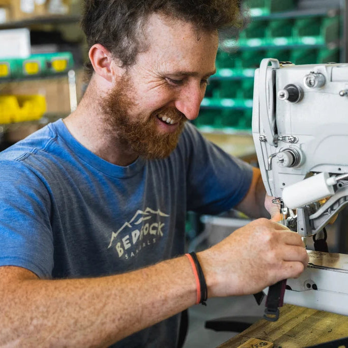 A smiling man with a beard wearing a blue t-shirt with a 'Bedrock Samples' logo, operating a white industrial sewing machine in a workshop setting.