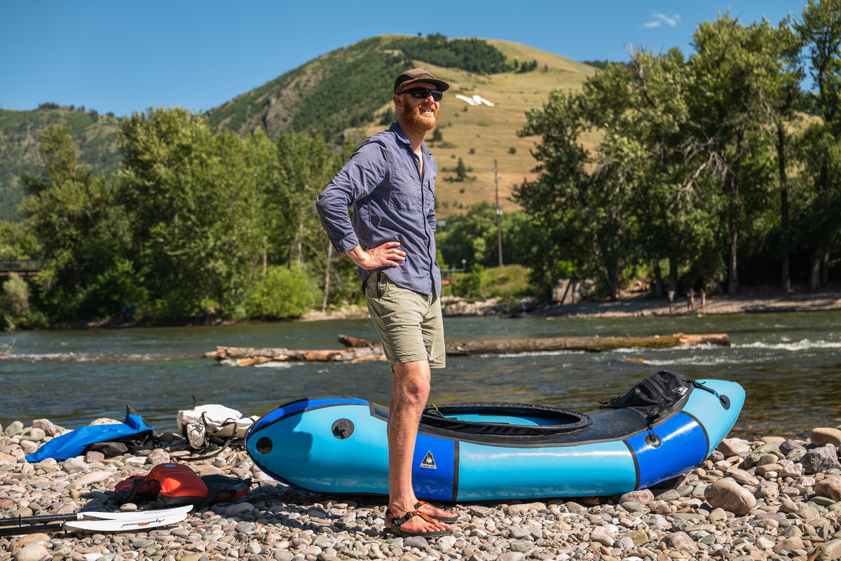 A man stands on a rocky riverbank wearing sunglasses, a blue button-up shirt, and khaki shorts, with a bright blue inflatable raft beside him and a river, trees, and a grassy mountain in the background.