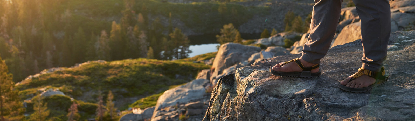 person wearing yellow cairn evo sandals with a closeup on their feet while they are standing on a rocky surface overlooking an alpine lake
