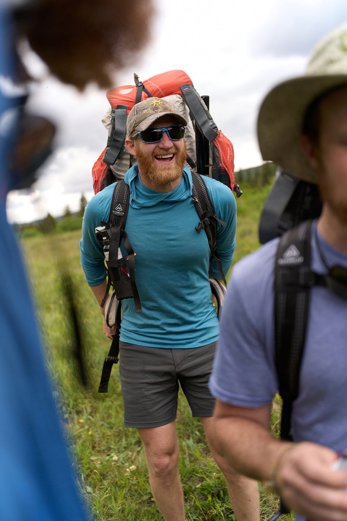 A man hiking outdoors wearing a large red and black backpack, a blue long-sleeve shirt, gray shorts, sunglasses, and a camouflage cap, with other people partially visible in the foreground.