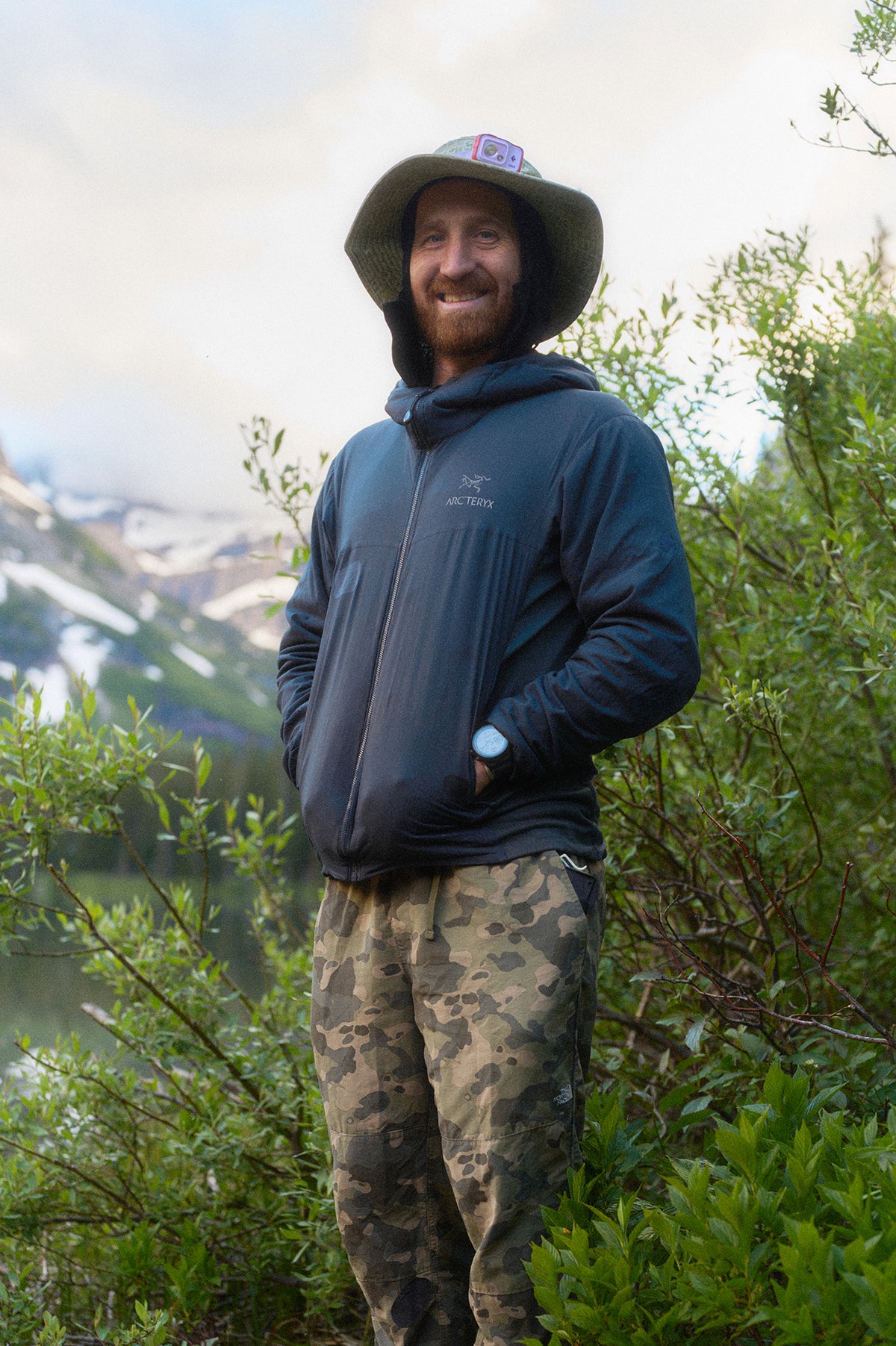 A smiling man wearing a dark blue Arc'teryx zip-up hoodie, camouflage pants, and a green bucket hat stands outdoors in front of green bushes and mountains.