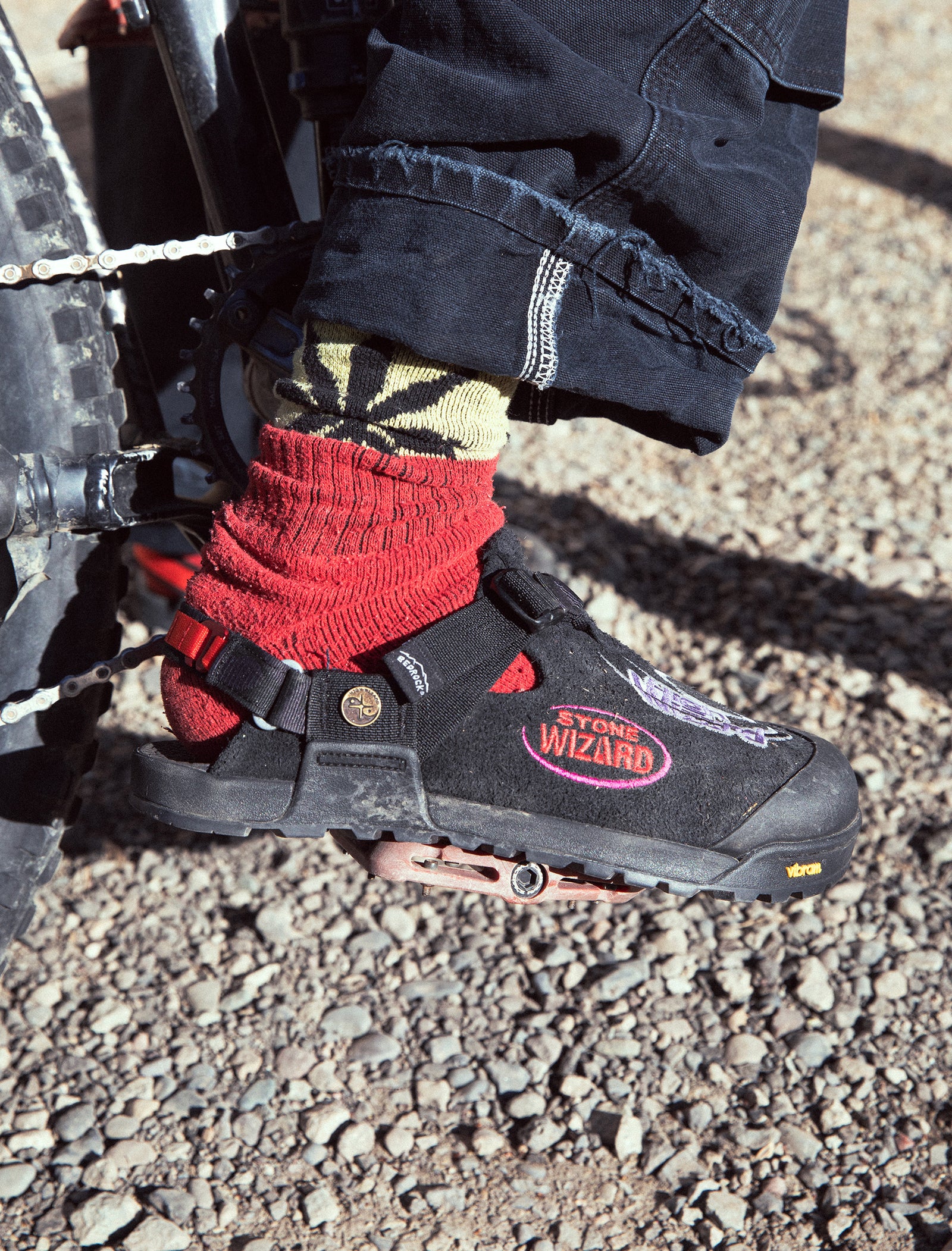 Person wearing mountain clogs with red accents and a visible brand logo, standing on a gravel surface.