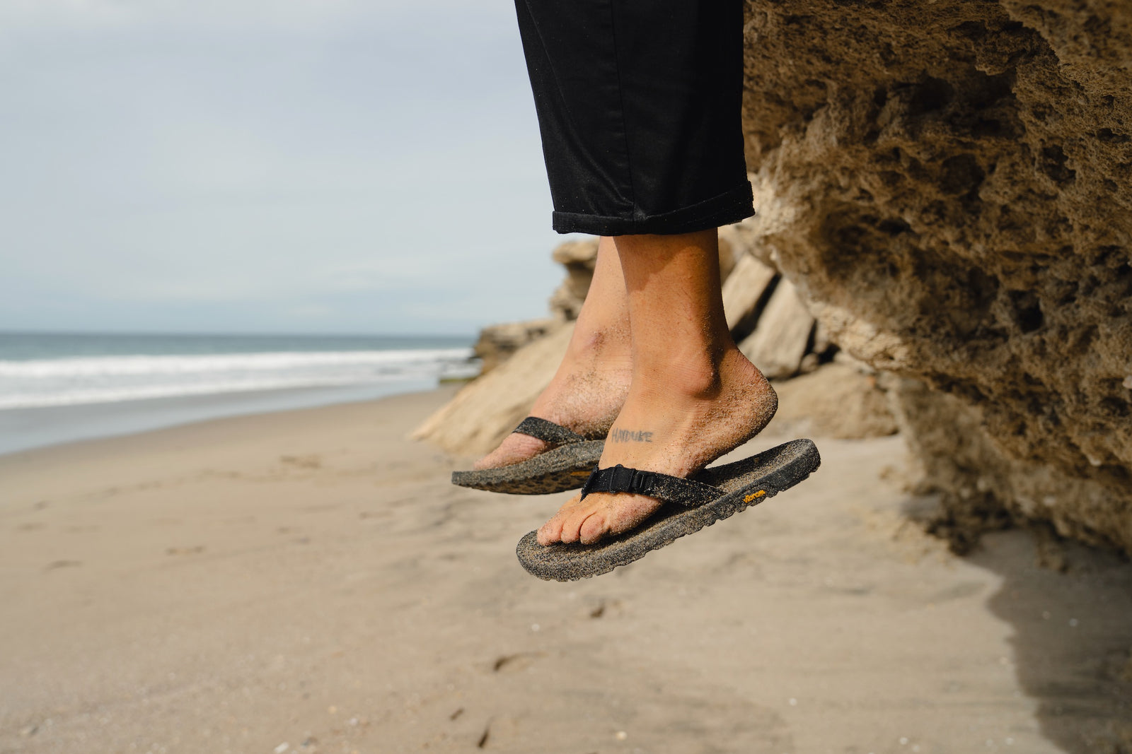 Person wearing black rockhound sandals on a beach with ocean and rocks in the background