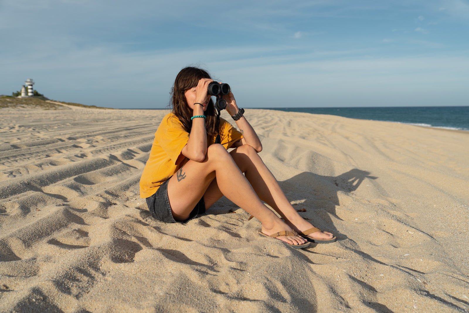 Person looking through binoculars on a sandy beach with a lighthouse in the background wearing nubuck rockhound sandals.