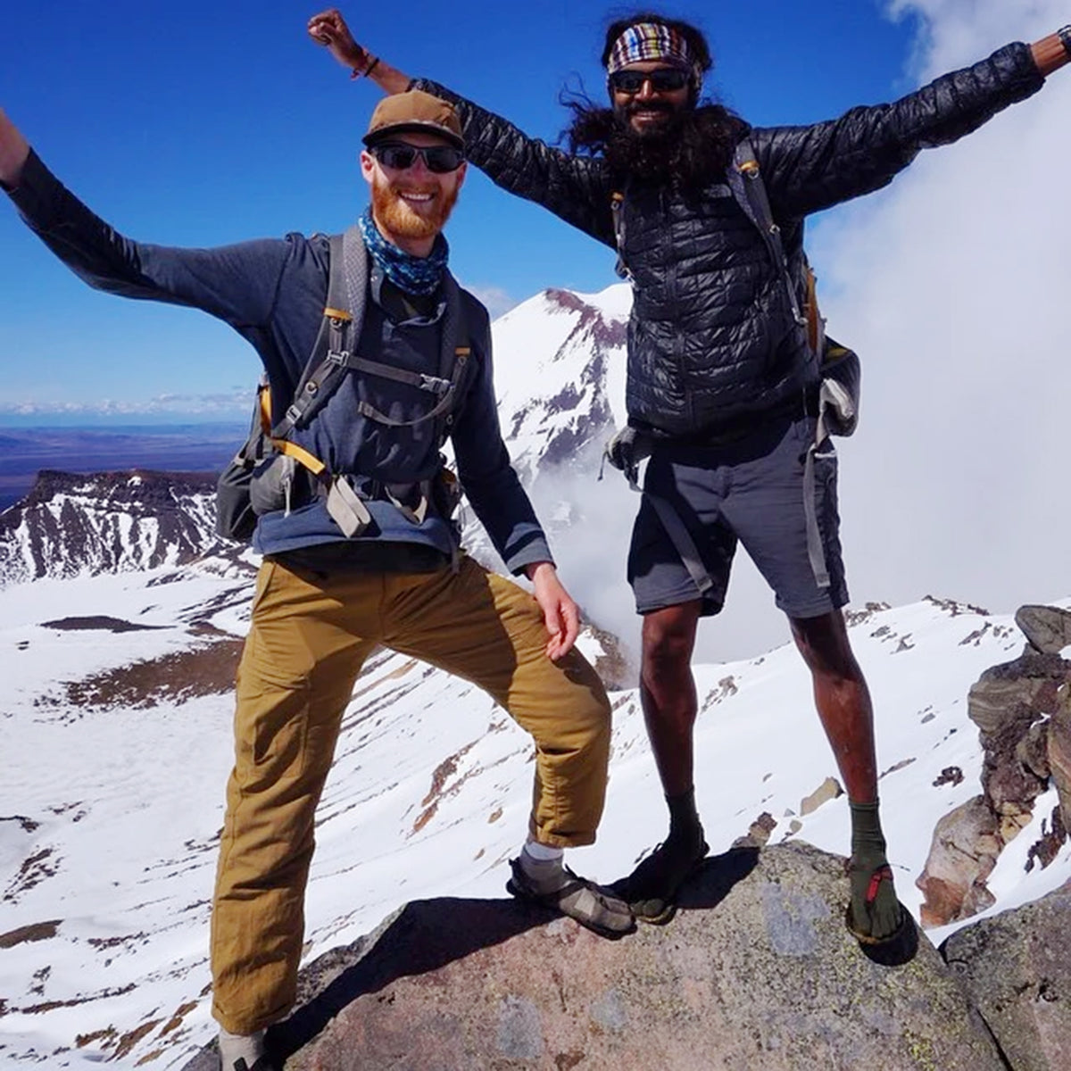 Two hikers posing on a snowy mountain peak with their arms raised in triumph under a bright blue sky.