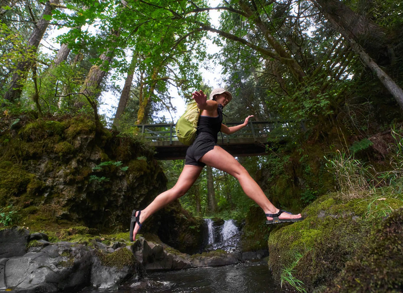 A woman in a black swimsuit jumps across a rocky stream in a mossy forest, carrying a light green backpack. A small wooden bridge and dense green trees are visible in the background.