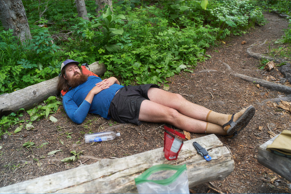A bearded man wearing a blue long-sleeve shirt, black shorts, with socks and sandals on his back on a log in a forest clearing, with a water bottle, red cup, and knife nearby.