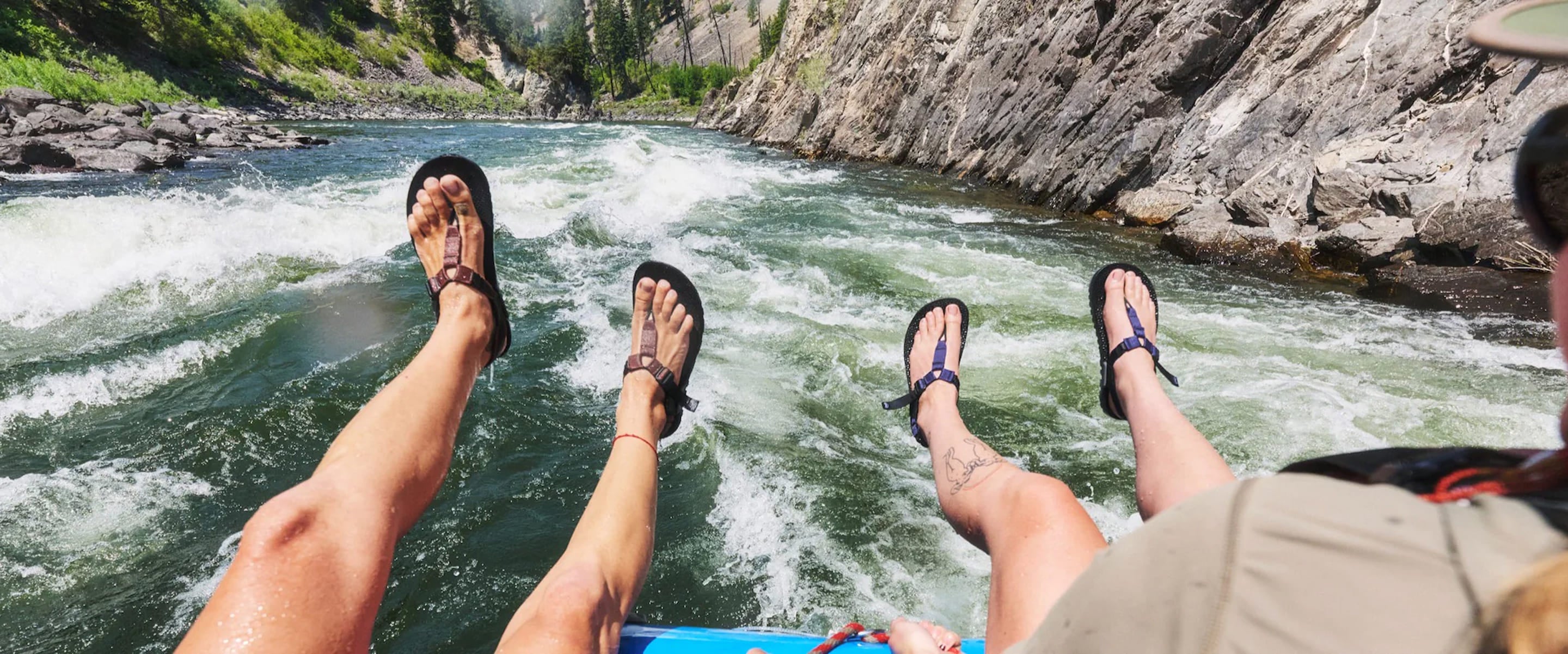 A first-person view from a raft on a fast-moving river, with two people's legs and feet in black flip-flops dangling over the side, surrounded by rocky canyon walls and green foliage.