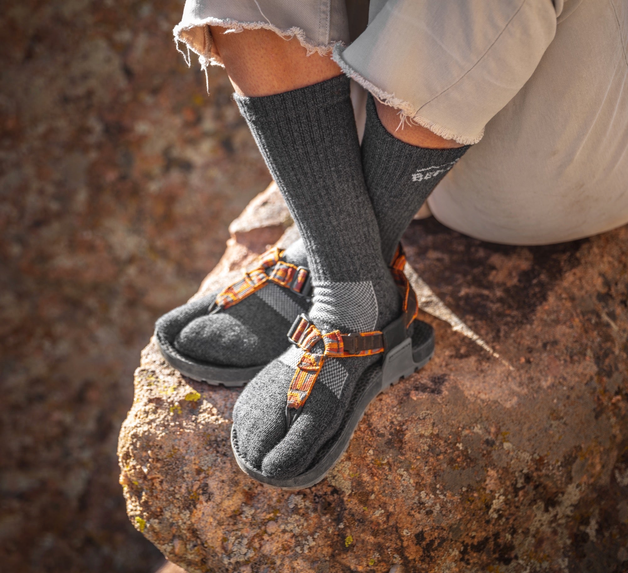 A person on a rock wearing a pair of cairn sandals with a pair of gray Bedrock x Injinji crew split-toe socks on. 